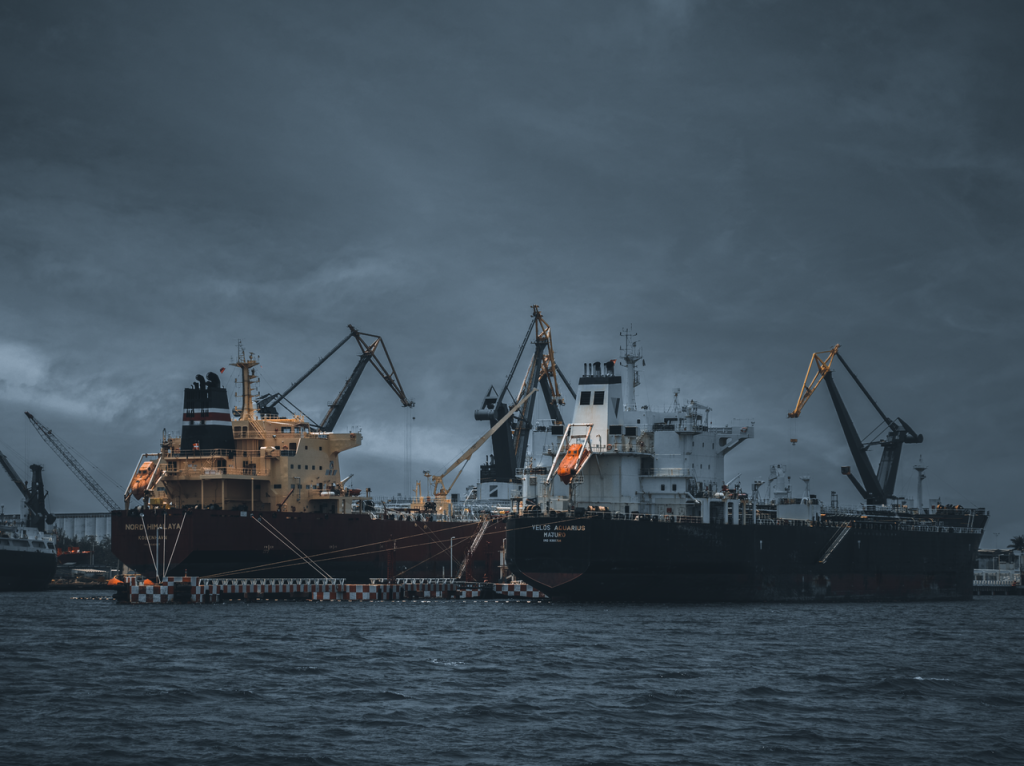 Cargo ships at a bustling port under cloudy skies, cranes ready for loading.